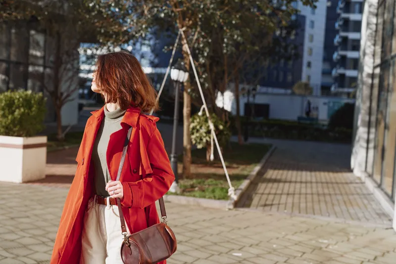 Young woman in red coat walks outdoors in sunny urban environment holding brown purse looking sideways with casual confident expression during daytime