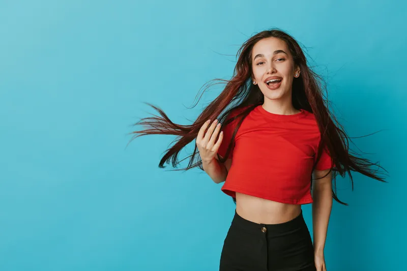 Young woman with long hair wearing red crop top and black pants standing against blue background smiling and holding hair with playful expression in studio portrait. Casual style and vibrant mood.