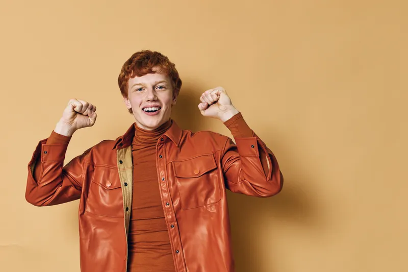 Young man with red hair smiling and celebrating success with raised fists in casual orange outfit against plain beige background. Positive expression and joyful mood.