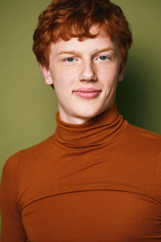 Portrait of young man with red hair wearing orange turtleneck against green background, looking at camera with neutral expression and natural lighting for studio shot.