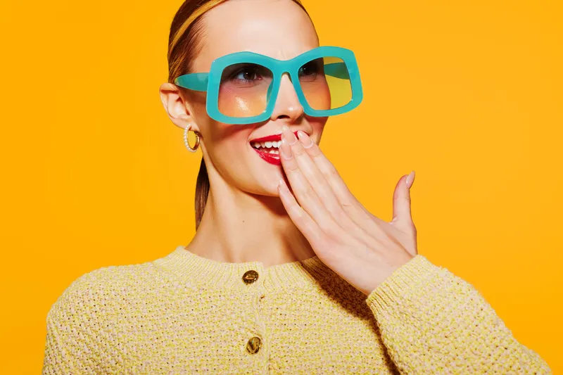 Stylish woman in oversized turquoise sunglasses and yellow sweater laughs covering mouth with hand. Bright orange background creates vibrant, playful mood. Concept for fashion, summer, and fun.