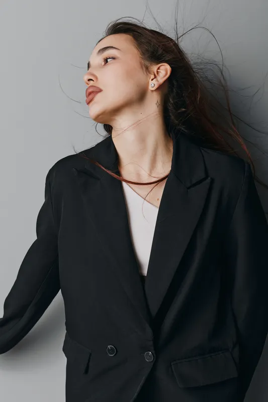 Young woman in black blazer with flowing hair looking sideways against gray background. Portrait of confident female model with natural makeup and earrings in studio lighting.