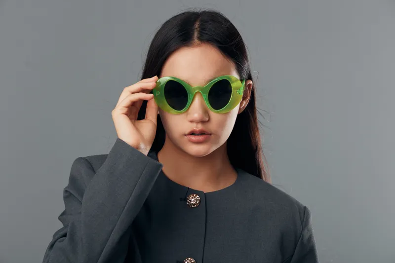 Woman with long hair wearing green sunglasses and gray jacket posing against gray background in studio. Stylish female model looking at camera with serious expression and fashion accessories.