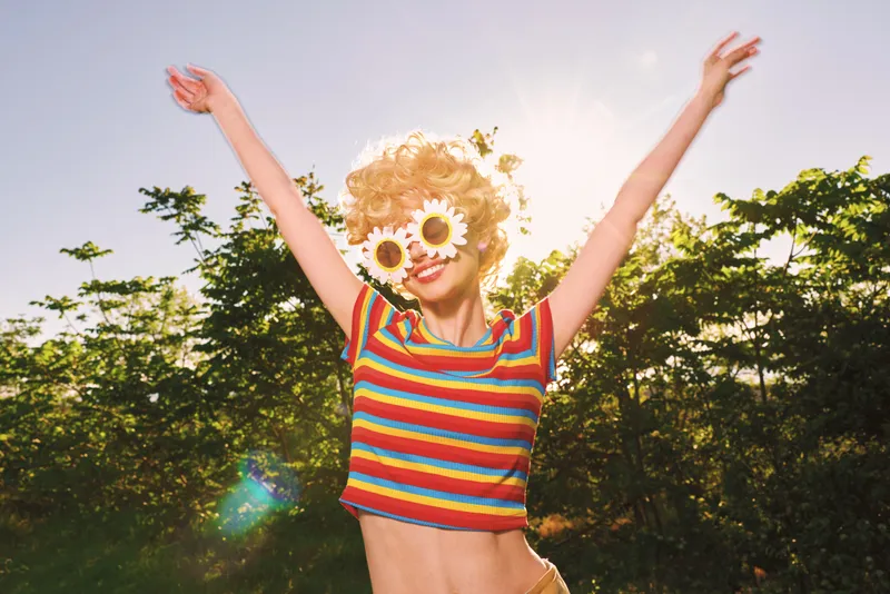 Young woman with sunflower sunglasses leaps in a sunlit field wearing a rainbow striped top, embodying carefree retro energy and warm summer happiness.