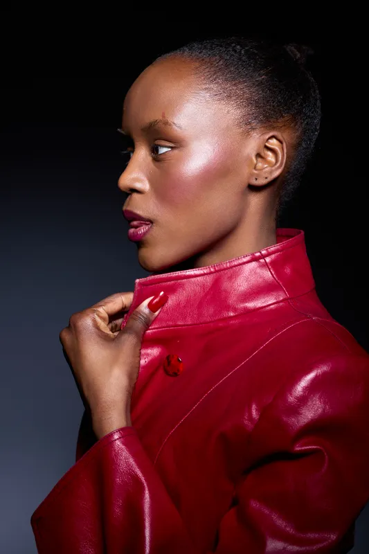 Profile of a dark skinned African woman wearing a red leather jacket. She looks to the side with a confident expression, showcasing smooth skin and elegant posture on a dark background.
