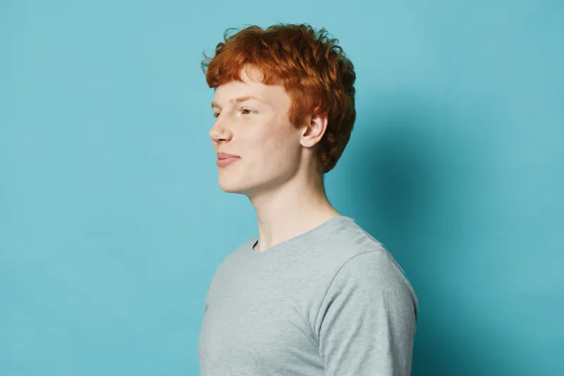Young man with red hair in gray shirt standing in profile against blue background with thoughtful expression and soft lighting for portrait or lifestyle concept.