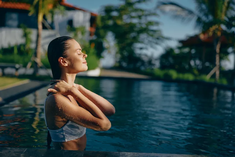Relaxed woman enjoys a sunlit poolside moment, standing in water with shoulders exposed, serene expression, chic resort atmosphere, outdoor summer lifestyle scene, tranquil leisure.