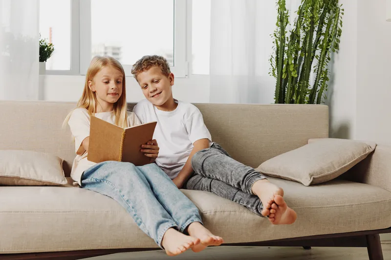 Two children sitting on beige couch reading a book together in bright living room, cozy home atmosphere and sibling bonding moment captured.