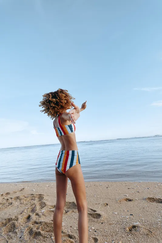 Woman standing in colorful swimsuit on sandy beach near calm ocean, summer vacation mood, sunny sky, outdoor sunlight, soft breeze, full length shot, relaxation and travel