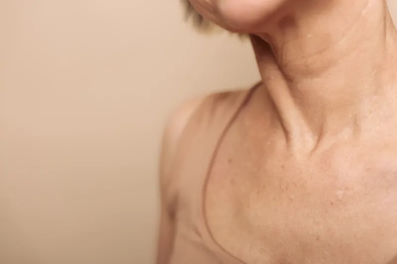 Close-up of mature female neck and shoulder with natural skin texture, wearing a beige tank top. Concept for aging, skin care, health, and natural beauty in soft lighting.