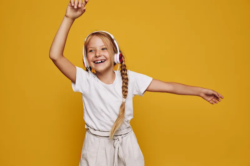 Happy girl with long blonde braid wearing headphones dances joyfully on a yellow background. She smiles widely with arms raised. Studio shot for music and fun concept.