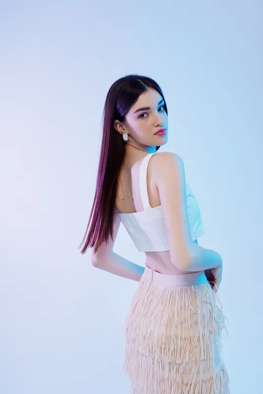 Young woman beauty fashion model poses in white top and fringed skirt on light background. Concept of style, elegance, and modern trends in female fashion.