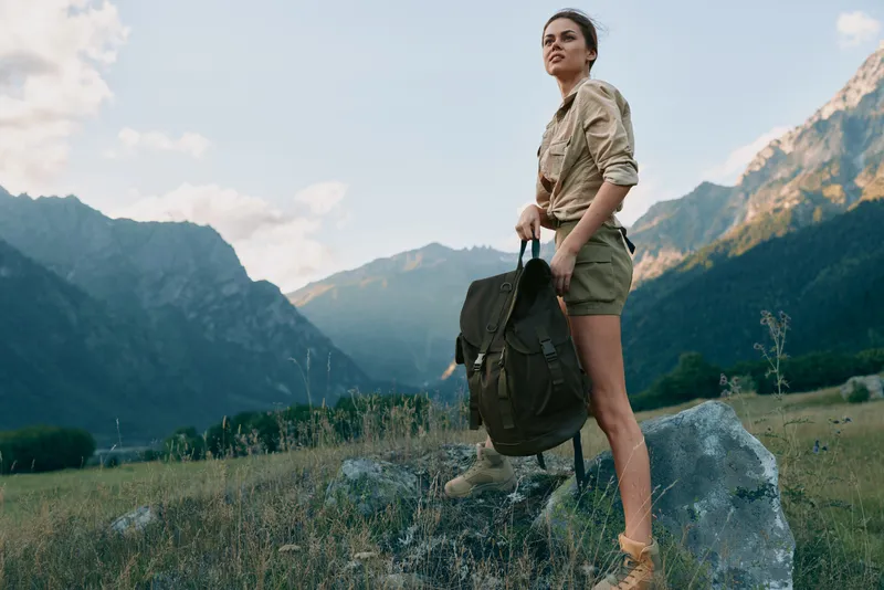 Young woman with long hair stands on a rocky meadow with a backpack, ready for a mountain hike in a vast natural landscape, conveying focus and anticipation