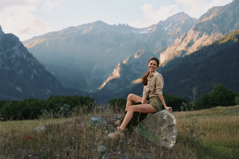 Young woman sitting on rock in mountain meadow, smiling relaxed, casual outfit, sunny day, natural landscape, outdoor lifestyle, scenic background