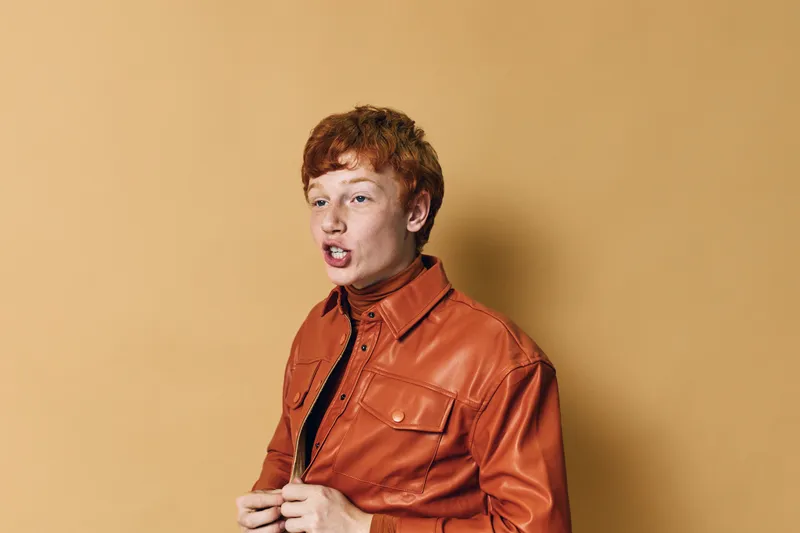 Young man with red hair wearing orange leather jacket stands against beige background, looking sideways with expressive face and casual posture indoors.