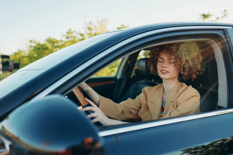 Cheerful woman driving a car outdoors, sunlit moment, relaxed posture, curly hair, tan jacket, soft breeze, natural colors, retro vibe, warm light, casual lifestyle with carefree mood.