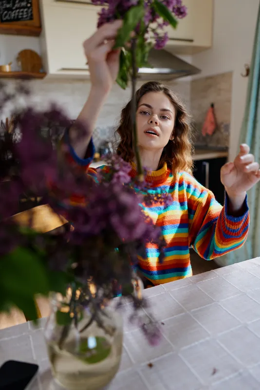 A woman in a bright striped sweater arranges flowers at a kitchen table, capturing a cozy home moment, emphasizing lifestyle, creativity, and warmth in a cheerful scene.