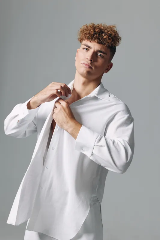 Young man with curly hair wearing white shirt posing against gray background, adjusting collar with serious expression and casual style in studio lighting.