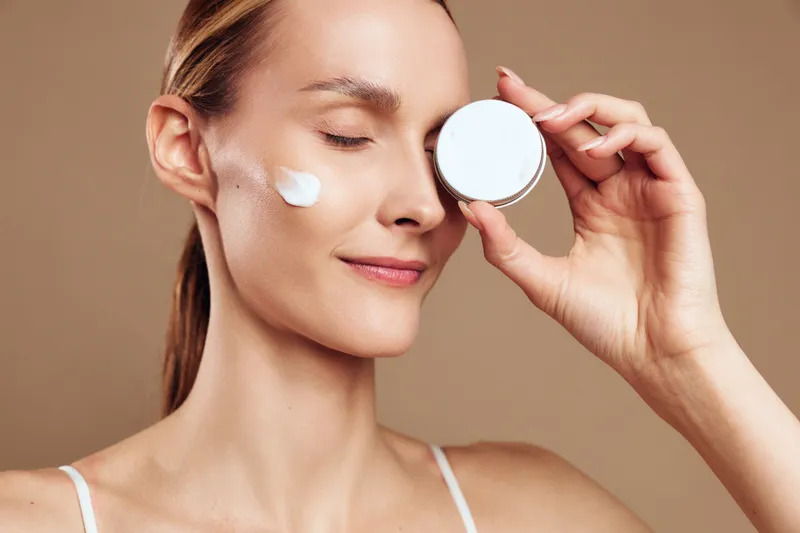Young woman applies face cream on cheek holds jar of skincare product close to face eyes closed enjoys beauty routine isolated on beige background with natural makeup.