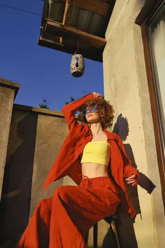 A stylish woman in a scarlet jumpsuit and yellow crop top poses on a sunlit terrace, shielding her eyes with one hand while striking a confident, relaxed pose