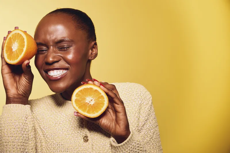 Happy African woman with dark skin smiling and holding two orange halves near her face on yellow background. Concept of healthy lifestyle, vitamin C, and natural beauty.