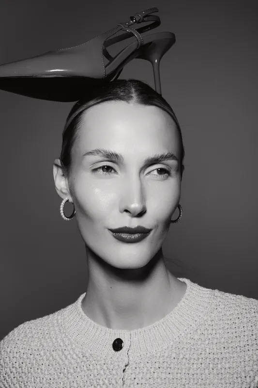 Caucasian woman with fair skin and slicked hair balances high heel shoe on head, wearing hoop earrings and knit sweater studio portrait.