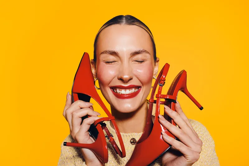 Happy woman with closed eyes holds red high heel shoes near face on yellow background. Concept of fashion, style, footwear, and joyful emotions in studio lighting.