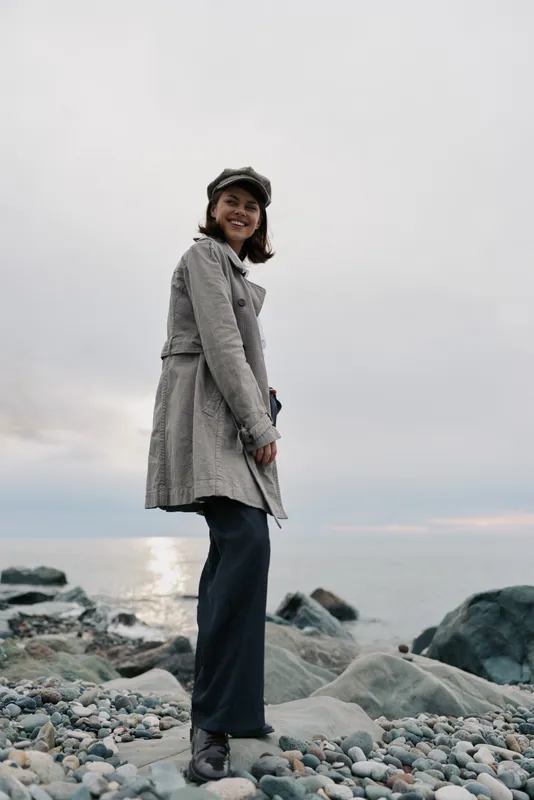 Smiling woman in stylish coat and hat stands on rocky beach at sunset. Casual autumn fashion concept with natural light and calm ocean background for outdoor lifestyle.