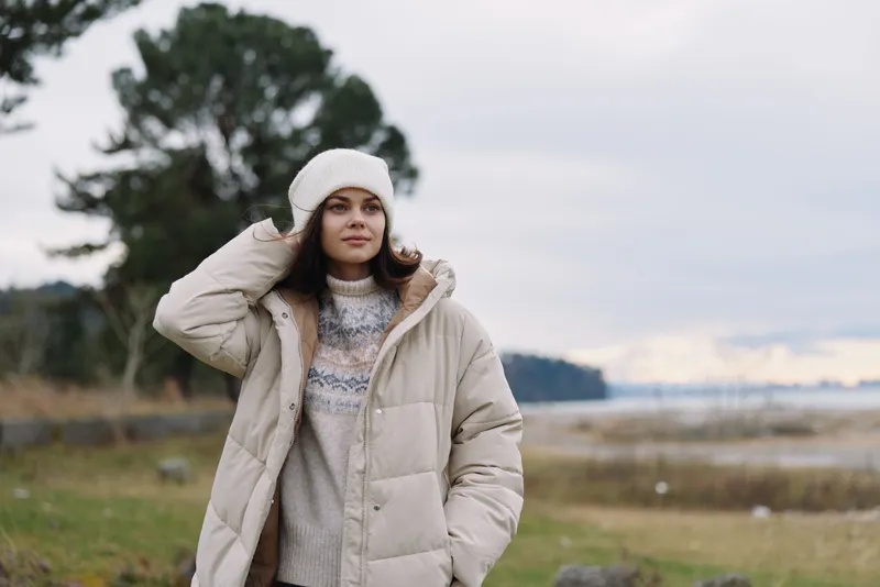 Woman in a beige puffer coat and knit sweater stands outdoors by a lake, wearing a beanie under a cloudy sky, capturing a calm winter landscape