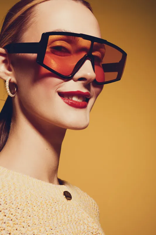 Stylish woman wearing oversized orange sunglasses and hoop earrings smiles confidently against a warm yellow background. Fashion portrait with trendy accessories and makeup.