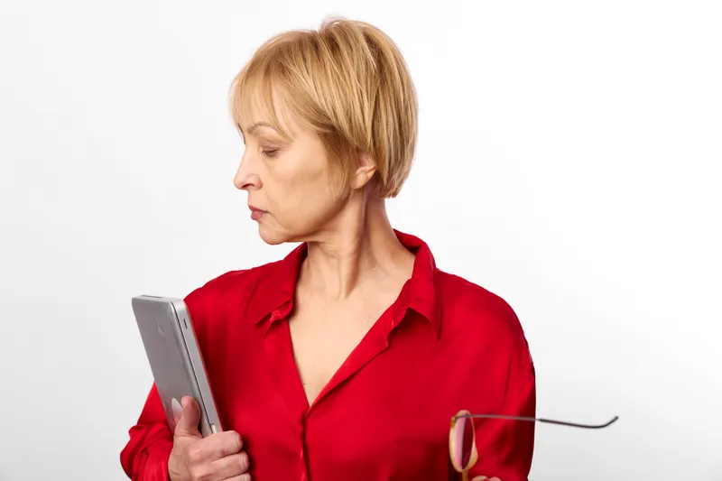 Woman portrait in red blouse holding a tablet, sipping wine from glass, calm adult female on plain white background, indoor setting, side view, copy space.