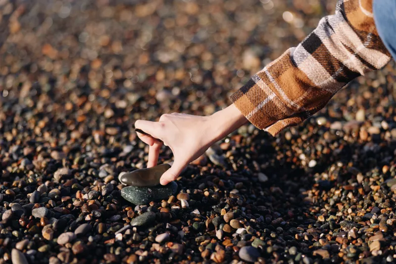 Hand reaching to pick a smooth pebble on a pebbled beach, outdoors, plaid sleeve in view, close texture of stones and sand, calm natural scene with tactile detail.