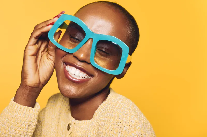 Happy African woman with dark skin wearing oversized blue glasses and a yellow sweater smiles with closed eyes on bright yellow background. Concept of joy, fashion, and beauty.