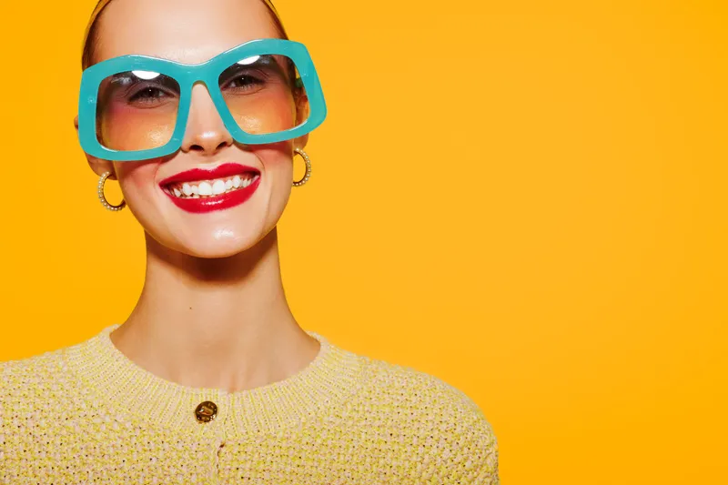 Smiling woman with bright red lipstick wears large blue sunglasses and yellow sweater against vibrant yellow background. Concept of summer fashion, style, and confident beauty portrait.