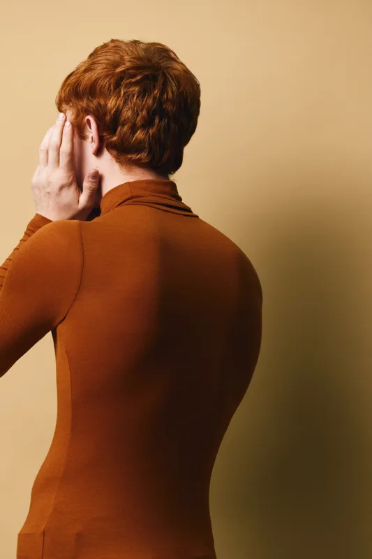 Man with red hair and brown turtleneck facing away, hand covering face, posing against beige background. Casual style with smooth skin and neat haircut.