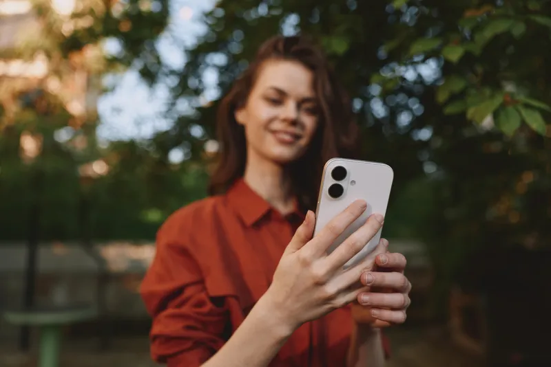 Smiling woman in a casual red shirt takes a selfie outdoors in a park with greenery. A lifestyle portrait capturing a moment of modern, everyday technology use and relaxed fashion