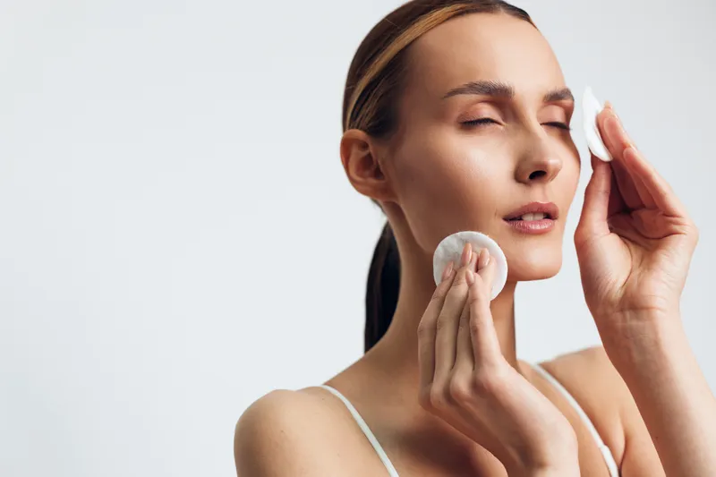 Young woman with closed eyes cleansing her face using cotton pads, clear skin care routine, gentle touch, natural light, minimalistic background, fresh healthy skin.