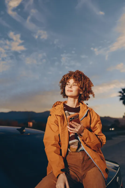 Woman lifestyle curly hair smartphone jacket outdoor sunset concept travel, young woman stands near car with mountains, holding phone, orange coat, warm evening light, golden sky, smile