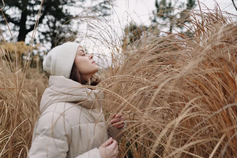Woman in a puffy coat and knit hat stands among tall dried grass in an open field, capturing a calm winter moment and a intimate connection with nature in a quiet outdoor scene.