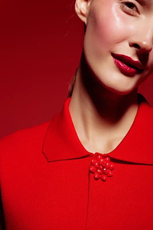 Close-up of a woman wearing a red textured top with a decorative clasp, red lipstick, and glowing skin. The vibrant red background enhances the elegant and stylish mood of the portrait.