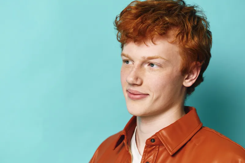 Redhead man looks sideways with smile wearing brown leather jacket on turquoise background. Youthful person with curly hair, casual style, and confident expression in closeup portrait with light.