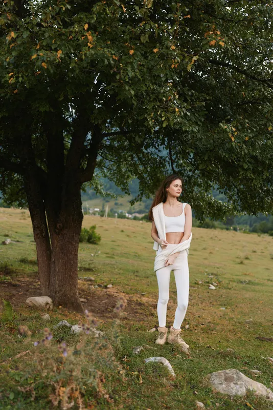 Young woman in white outfit stands near large tree in open field with mountains in background enjoying peaceful nature and relaxed lifestyle outdoors.