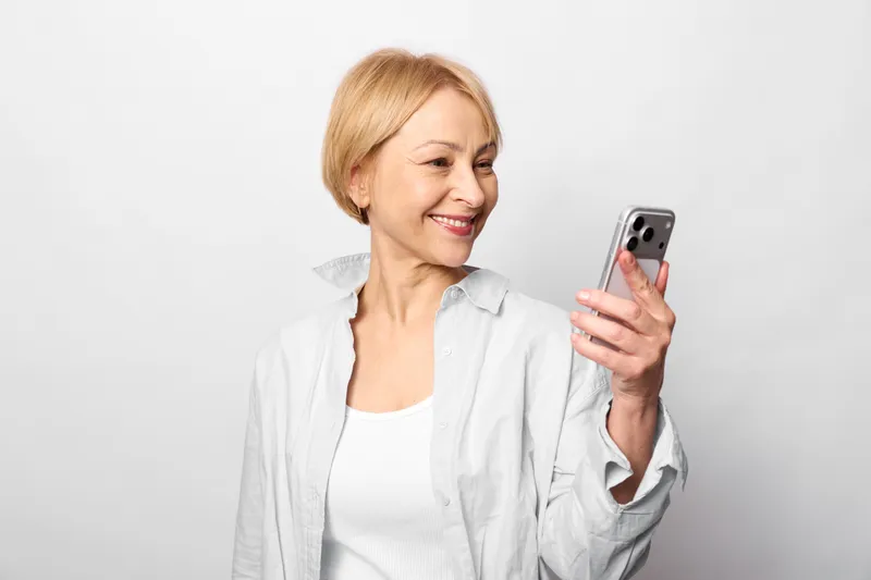 Woman smiling with smartphone, studio portrait on gray background, confident relaxed posture, light makeup, white shirt, looking at screen, friendly mood, copy space, today