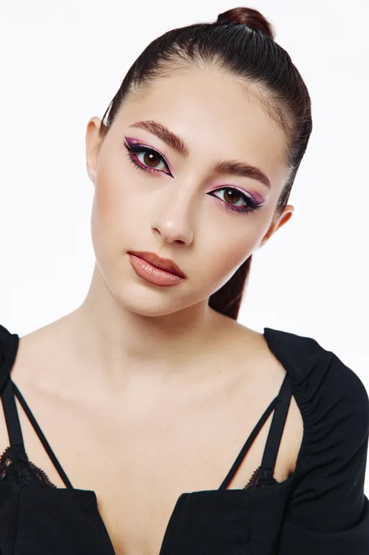 Young woman with smooth skin and creative pink eye makeup poses in a black top against white background. The image emphasizes beauty, fashion, and artistic makeup design.