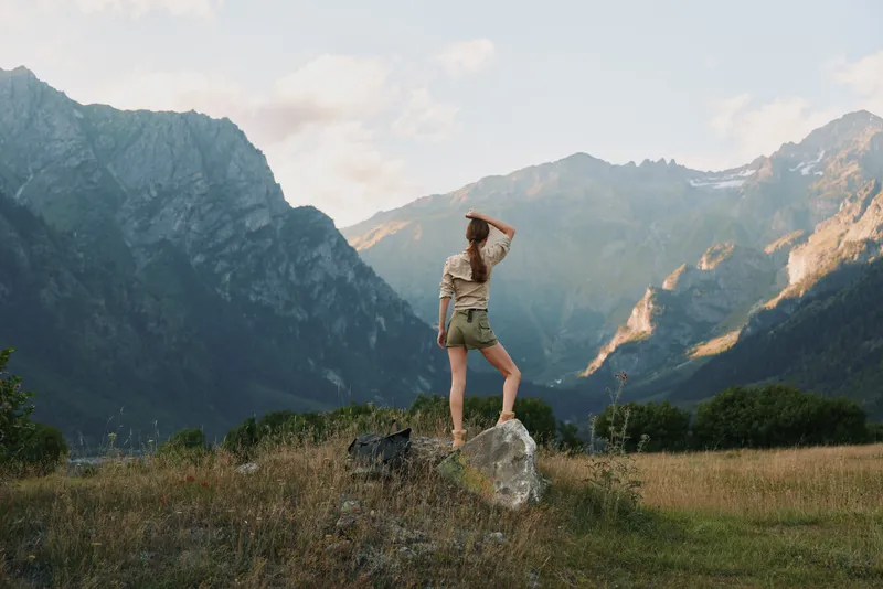 Woman standing on a rock in a mountain valley wearing a hat and casual outfit. Outdoor scenery with expansive landscape and sunny mood evokes adventure and freedom.