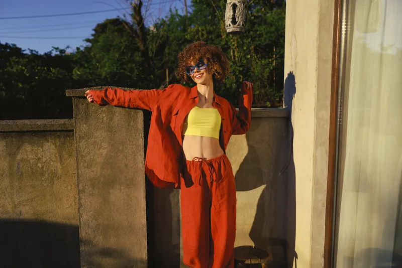Cheerful woman outdoors, bright yellow crop top, red suit, arms outstretched, sunlit balcony scene, warm natural tones, relaxed lifestyle, candid moment