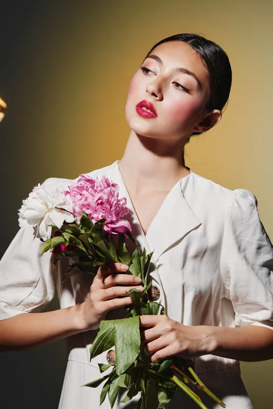 Elegant young woman with dramatic makeup holding fresh flowers in white blazer posing against gradient yellow background. Creative beauty and fashion portrait with styled hair and strong color