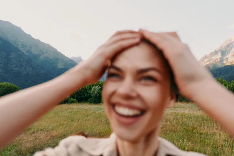 Young woman with short hair smiles joyfully while holding her head outdoors in a mountainous landscape during a bright day.