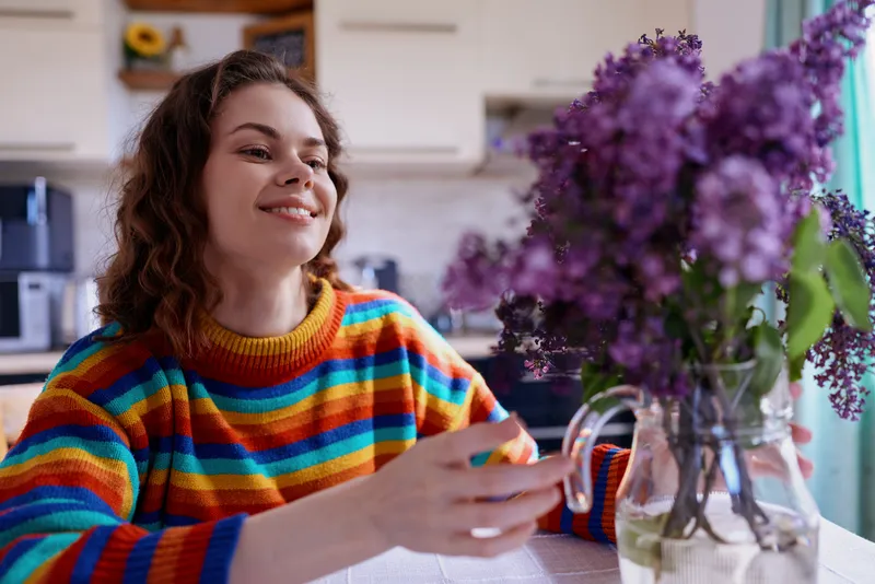 Cheerful woman indoors wearing a rainbow striped sweater, enjoying a bright home moment, flowers on the table, relaxed posture, warm light, casual lifestyle, cozy ambiance