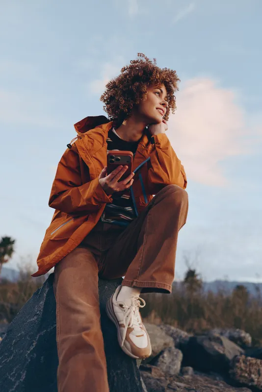 Woman with curly hair in an orange jacket holding a smartphone outdoors on rocky landscape, lifestyle travel scene at sunset sky, casual hiking mood, soft natural light, today